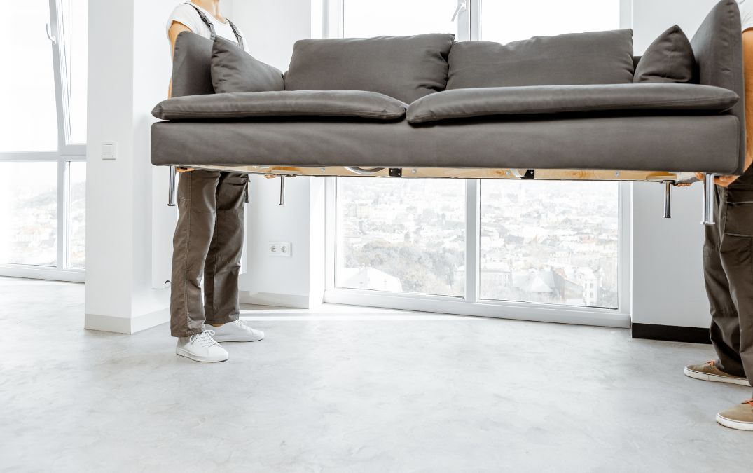 Men picking up a sofa and moving it across a concrete floor.